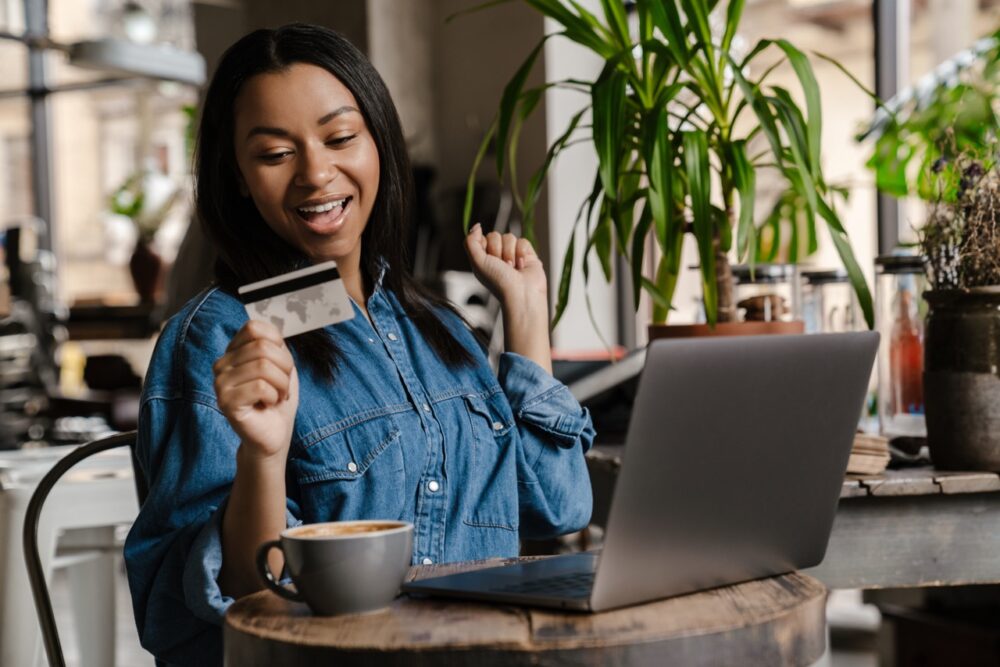 Happy woman holding credit card while shopping via laptop computer, sitting at the cafe indoors