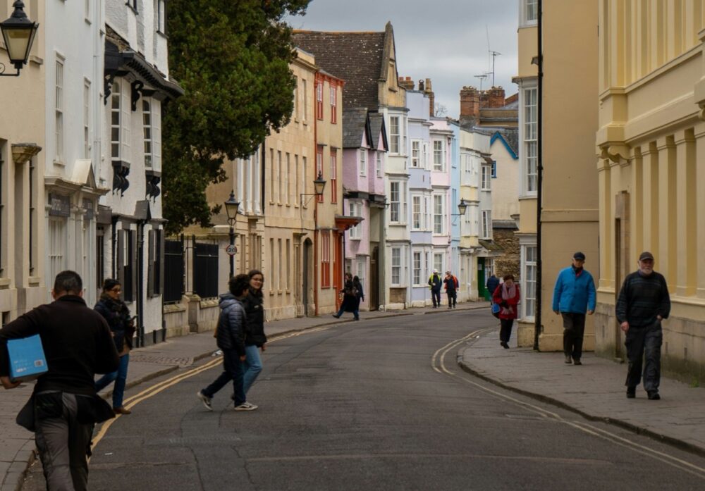 Shoppers walking through a busy British town centre lined with shops and upper floor buildings.
