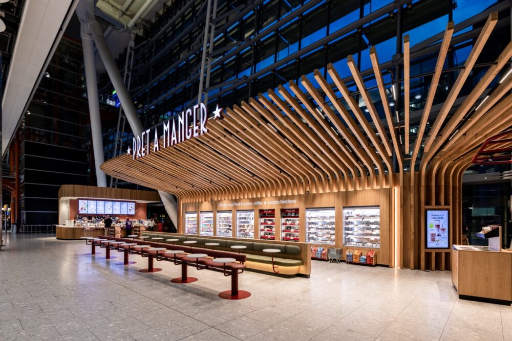 Pret A Manger’s new Heathrow Terminal 5 Arrivals store, designed by Draw Creative, uses a sweeping timber structure to frame the shopfront, food display and seating areas. The architectural treatment responds to the scale of the terminal, with illuminated branding, integrated lighting and a warm material palette creating a distinctive presence within the arrivals hall.