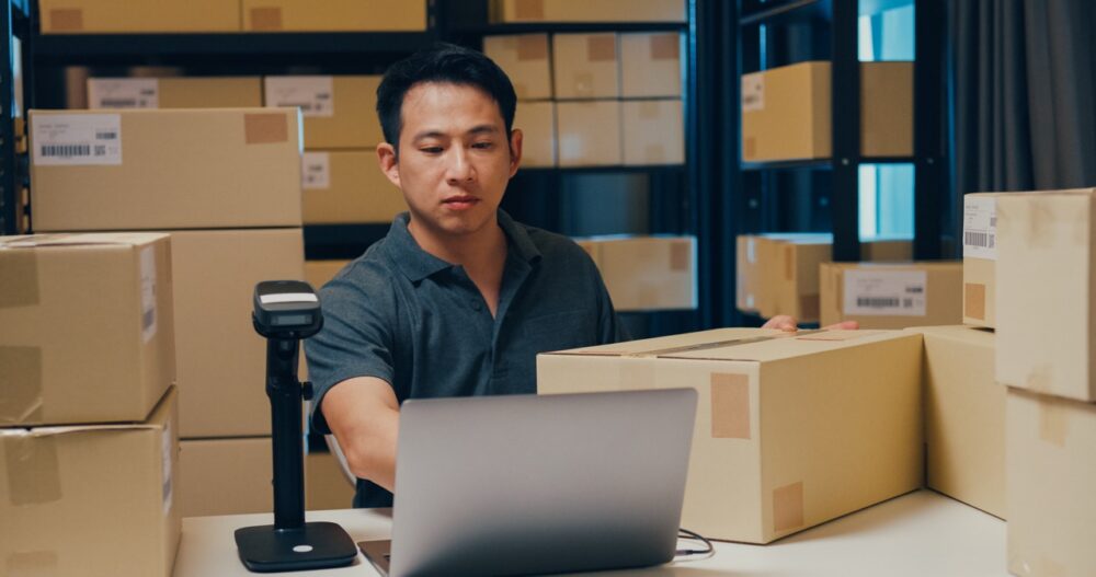 Asian businessman use barcode machine scan customer data in paper box and type register online information order detail in laptop for send express delivery at warehouse. Startup business concept.