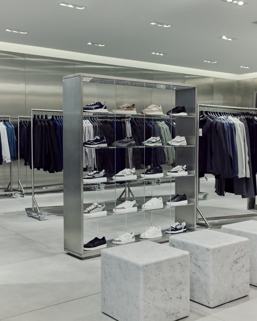 Inside the ARNE pop-up at Bluewater, trainers on acrylic shelves in a brushed steel frame, with clothing rails and marble cube seats against steel-clad walls