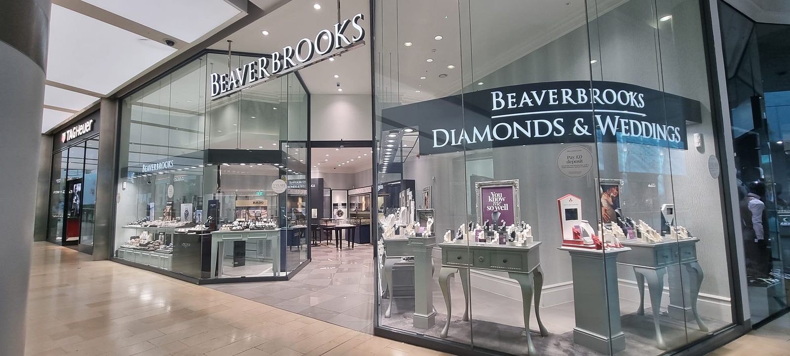 Beaverbrooks store frontage in a shopping centre, with illuminated signage, a Diamonds & Weddings window of jewellery displays and a TAG Heuer store next door