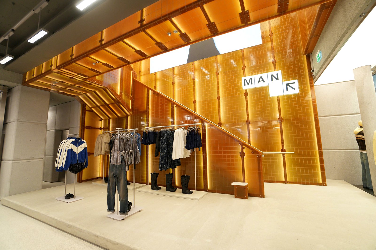 Staircase clad in amber-lit panels with MAN signage at Bershka Westgate Oxford, with rails of clothing and boots in front