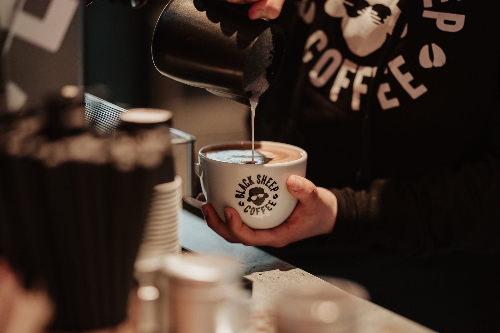 Barista pouring steamed milk into a Black Sheep Coffee branded cup at the counter