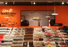 The Live @ bodycare content studio at Bodycare Sheffield, seen through glass with a camera on a tripod, behind tiered make-up displays against an orange wall