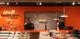 The Live @ bodycare content studio at Bodycare Sheffield, seen through glass with a camera on a tripod, behind tiered make-up displays against an orange wall