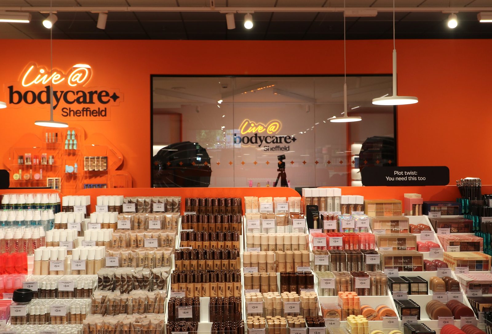 The Live @ bodycare content studio at Bodycare Sheffield, seen through glass with a camera on a tripod, behind tiered make-up displays against an orange wall