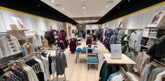 Interior of the Crew Clothing store at Westfield London, with knitwear, shirts and jackets on wall bays and rails either side of a central aisle