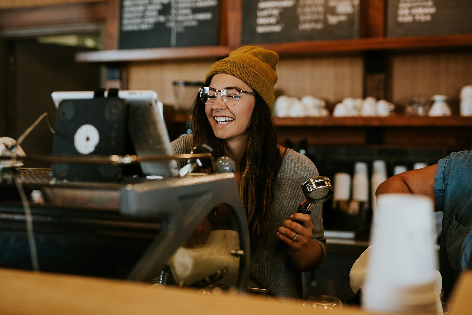 Smiling barista in a mustard beanie holding a portafilter behind the counter of a coffee shop