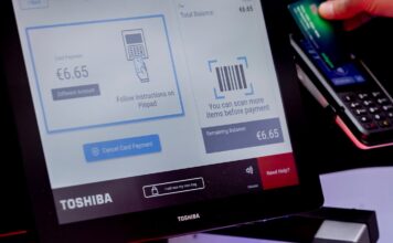 Self-checkout with a Toshiba screen showing a card payment, a red-lit barcode scanner and a shopper holding a card at the payment terminal