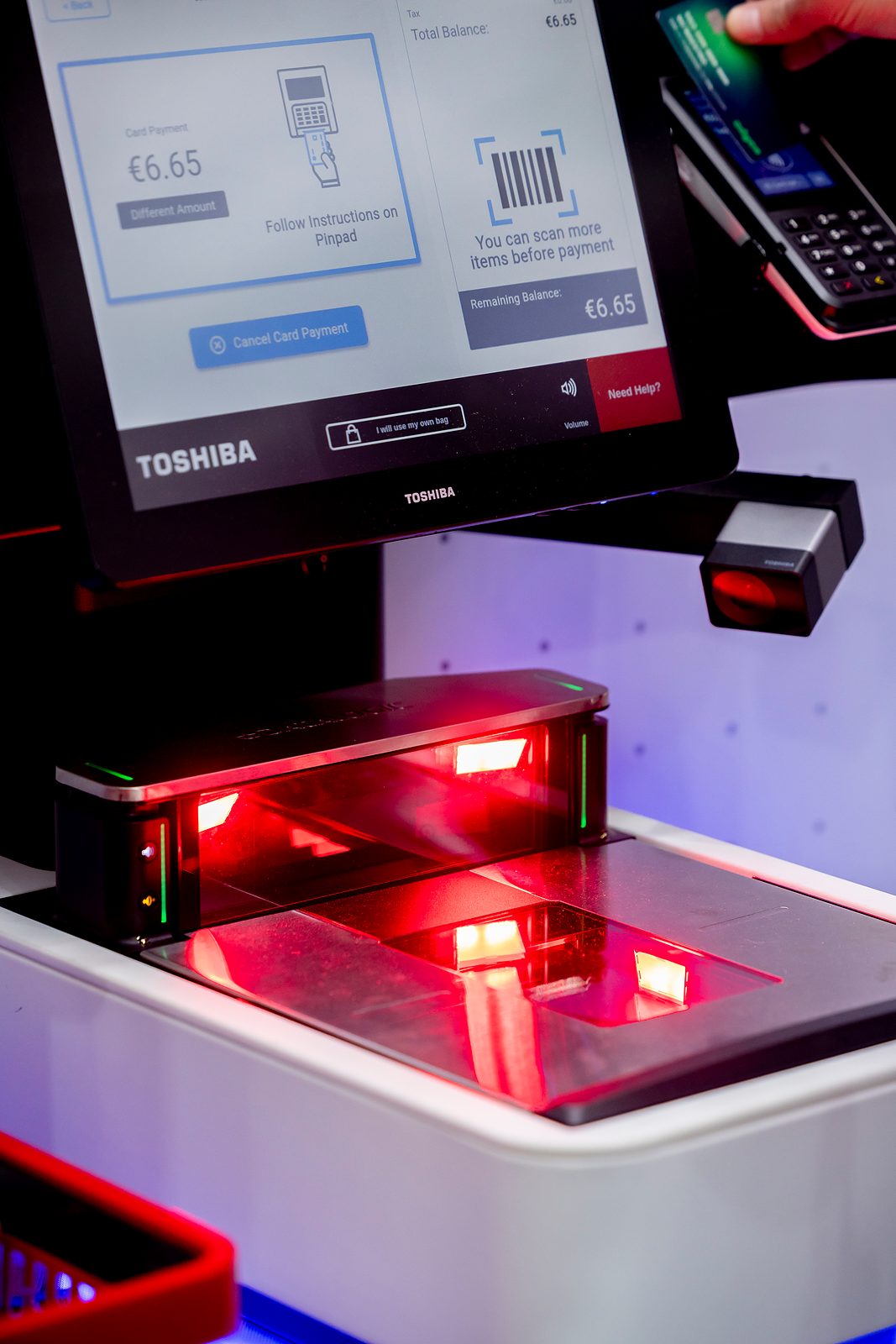 Self-checkout with a Toshiba screen showing a card payment, a red-lit barcode scanner and a shopper holding a card at the payment terminal