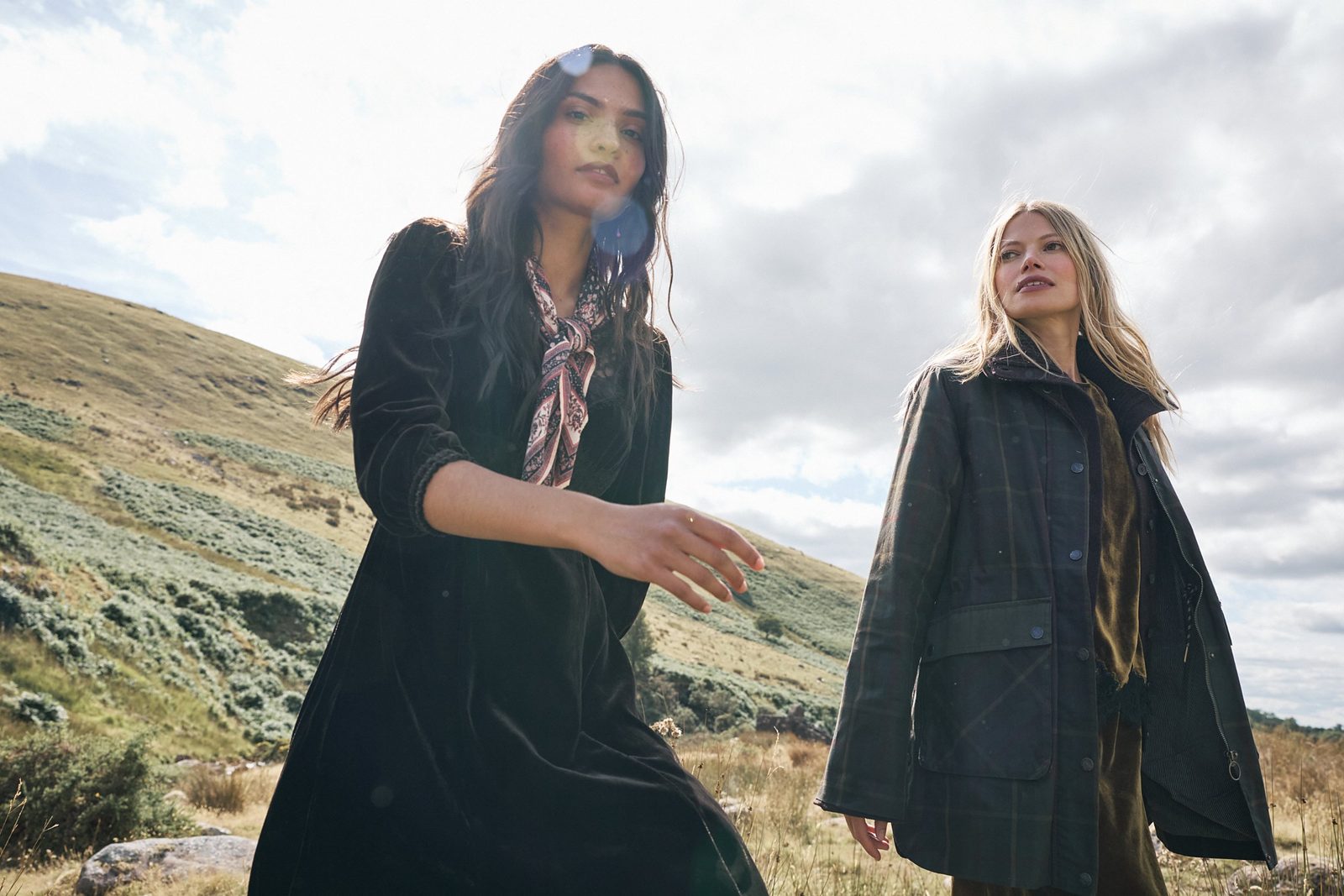 Two models outdoors in FatFace winter clothing, one in a black velvet dress and printed scarf, the other in a checked waxed jacket