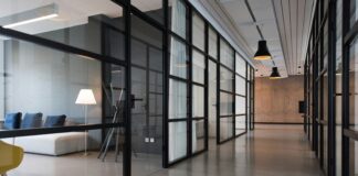 Modern office corridor lined with glass-walled meeting rooms, pendant lights and a lounge area