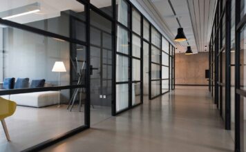Modern office corridor lined with glass-walled meeting rooms, pendant lights and a lounge area