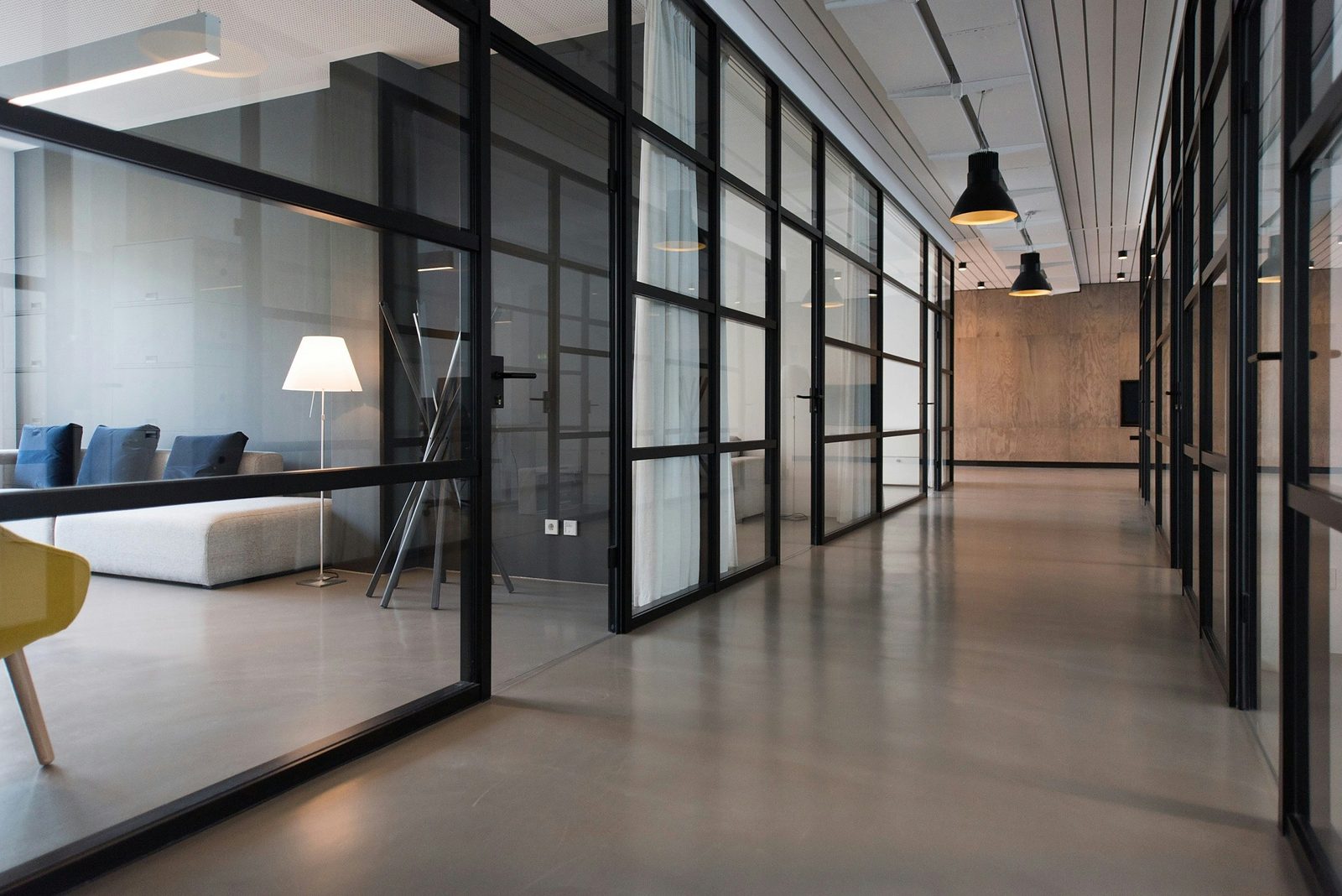 Modern office corridor lined with glass-walled meeting rooms, pendant lights and a lounge area
