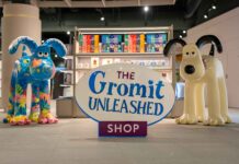 The Gromit Unleashed Shop sign between two painted Gromit sculptures, in front of shelving with Gromit Unleashed gift bags and mugs