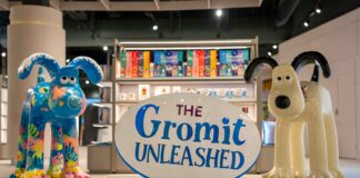 The Gromit Unleashed Shop sign between two painted Gromit sculptures, in front of shelving with Gromit Unleashed gift bags and mugs