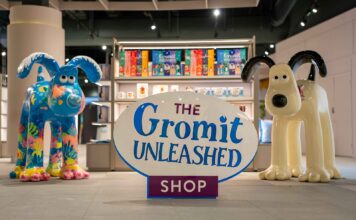 The Gromit Unleashed Shop sign between two painted Gromit sculptures, in front of shelving with Gromit Unleashed gift bags and mugs