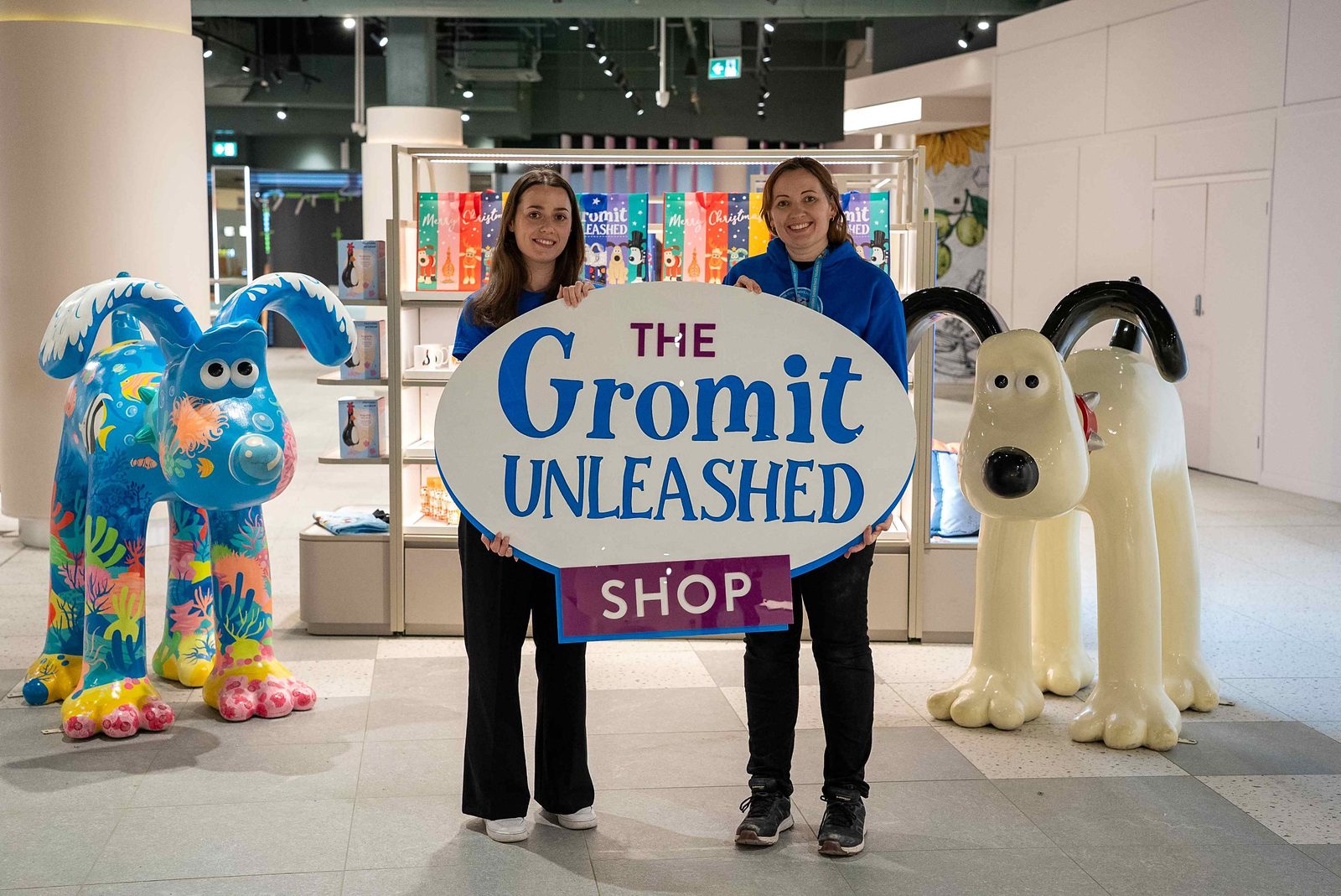 Two Gromit Unleashed team members holding The Gromit Unleashed Shop sign between two Gromit sculptures