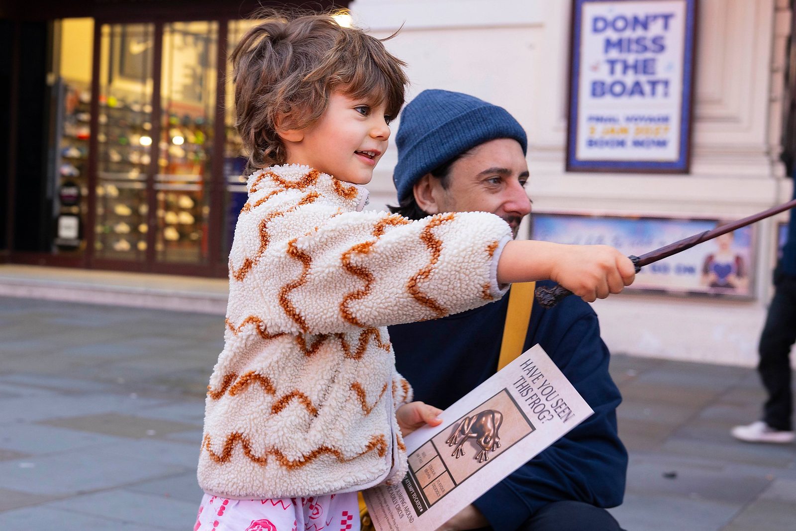 Child holding a toy wand and a 'Have you seen this frog?' flyer, with an adult crouching beside them on a London street