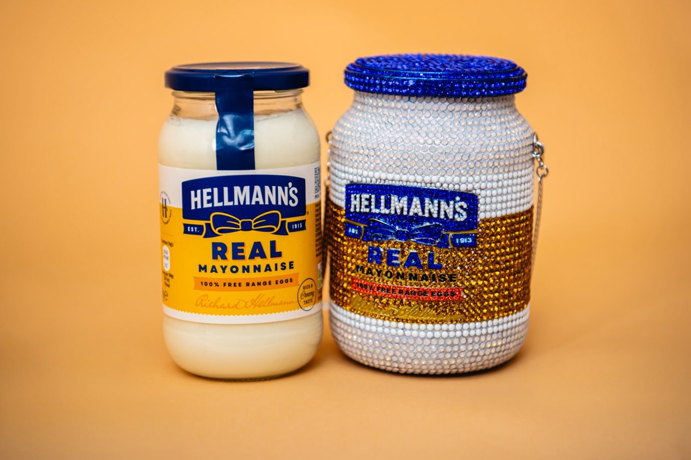 A jar of Hellmann's Real Mayonnaise beside Maya Grisham's crystal-covered handbag in the shape of the jar, with a blue crystal lid and a chain strap, on an orange background