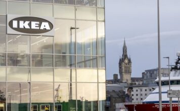 IKEA sign on the glass corner of a building in Aberdeen, with the harbour, a ship and a clock tower in the background