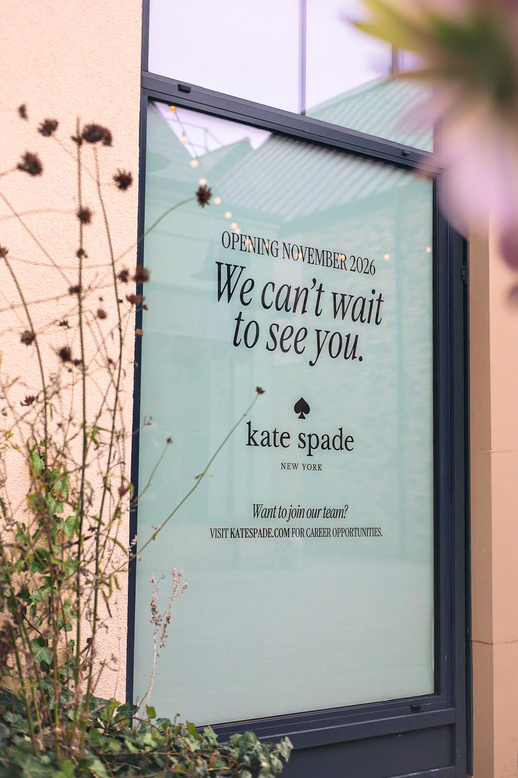 kate spade new york window graphic reading Opening November 2026 and We can’t wait to see you, with planting in front of the unit