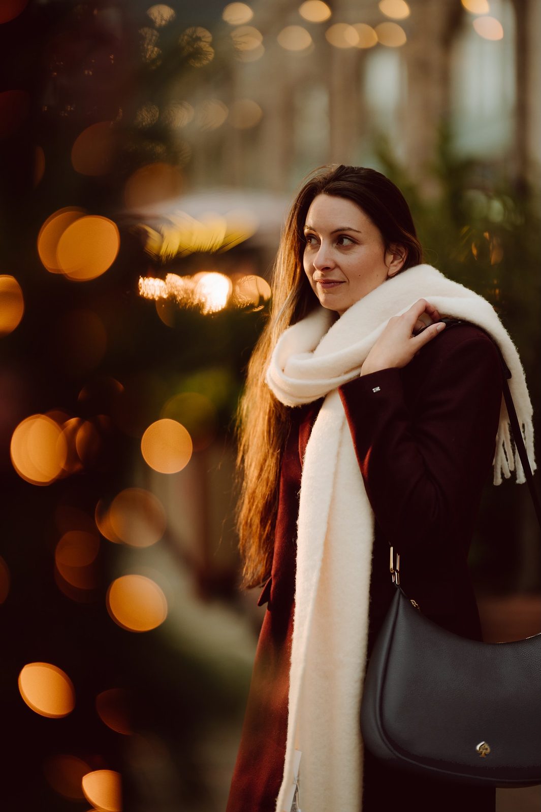 Woman in a burgundy coat and cream scarf carrying a black kate spade new york shoulder bag, with warm festive lights behind