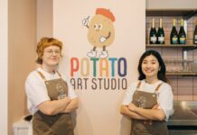 Two Potato Art Studio staff in branded aprons standing either side of the Potato Art Studio logo on a pale pink wall