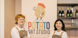 Two Potato Art Studio staff in branded aprons standing either side of the Potato Art Studio logo on a pale pink wall