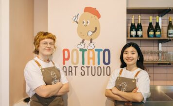 Two Potato Art Studio staff in branded aprons standing either side of the Potato Art Studio logo on a pale pink wall