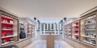 Room at Leica Store London Mayfair lined with red-backed glass cabinets of Leica cameras and lenses, with a central display plinth, bay windows and a marble table