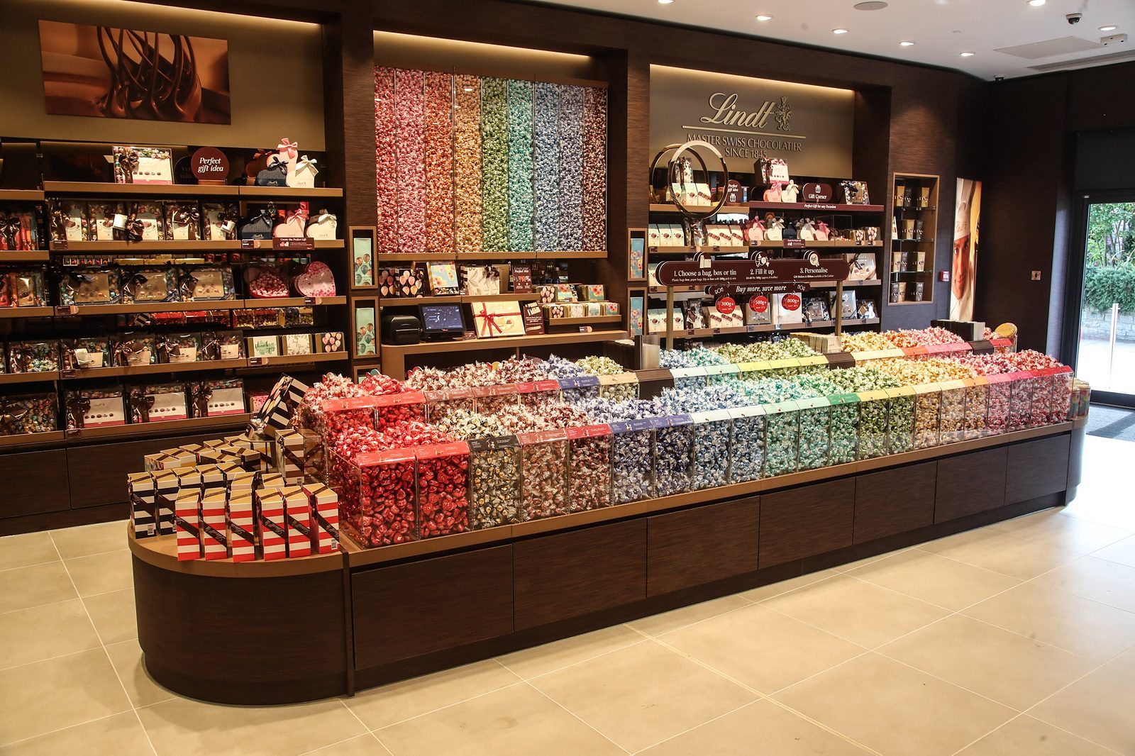 Angled view of a Lindt Chocolate Shop Pick & Mix counter, with gift boxes on dark wood shelving and a Lindt Master Swiss Chocolatier sign