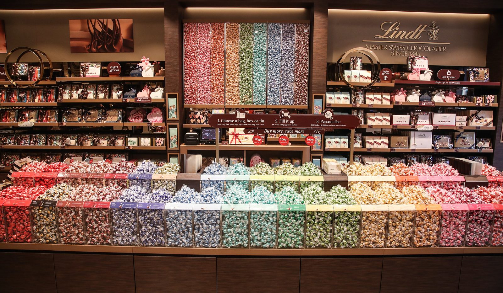 Lindt Pick & Mix counter with clear bins of LINDOR truffles in rainbow colours beneath a sign explaining how to choose, fill and personalise a bag
