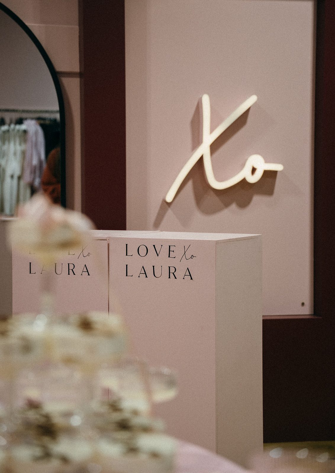 Illuminated Xo script sign on a pink wall above display plinths printed with the Love Laura Xo logo