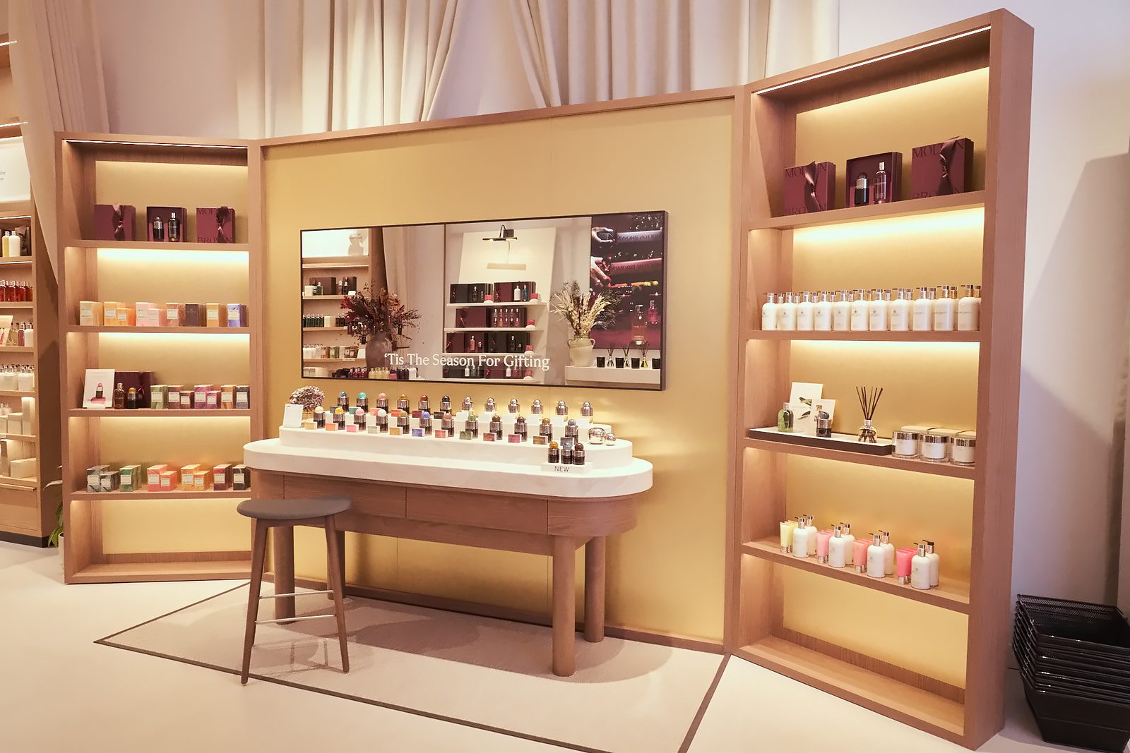 Fragrance bottles on an oval table in front of illuminated timber shelving and a screen showing a gifting campaign