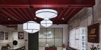 Lounge at New Balance's Arclight brand hub with leather sofas, a coffee table, a glass display case and a wooden New Balance sign on an exposed brick wall beneath paper pendant lights