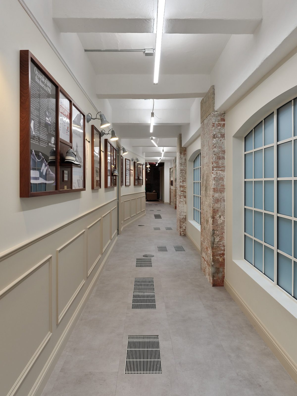 Long corridor with panelled walls, framed New Balance heritage displays, wall lamps and arched windows with exposed brickwork