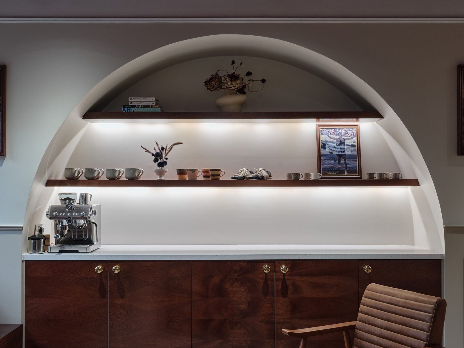 Arched alcove with lit wooden shelves holding mugs, trainers, flowers and a framed photograph above a coffee machine and wooden cabinets