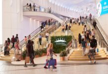 Shoppers on the gold staircase and escalators inside Outlet Shopping at The O2, with the outlet’s sign mounted on a green plant wall