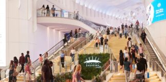 Shoppers on the gold staircase and escalators inside Outlet Shopping at The O2, with the outlet’s sign mounted on a green plant wall