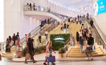 Shoppers on the gold staircase and escalators inside Outlet Shopping at The O2, with the outlet’s sign mounted on a green plant wall