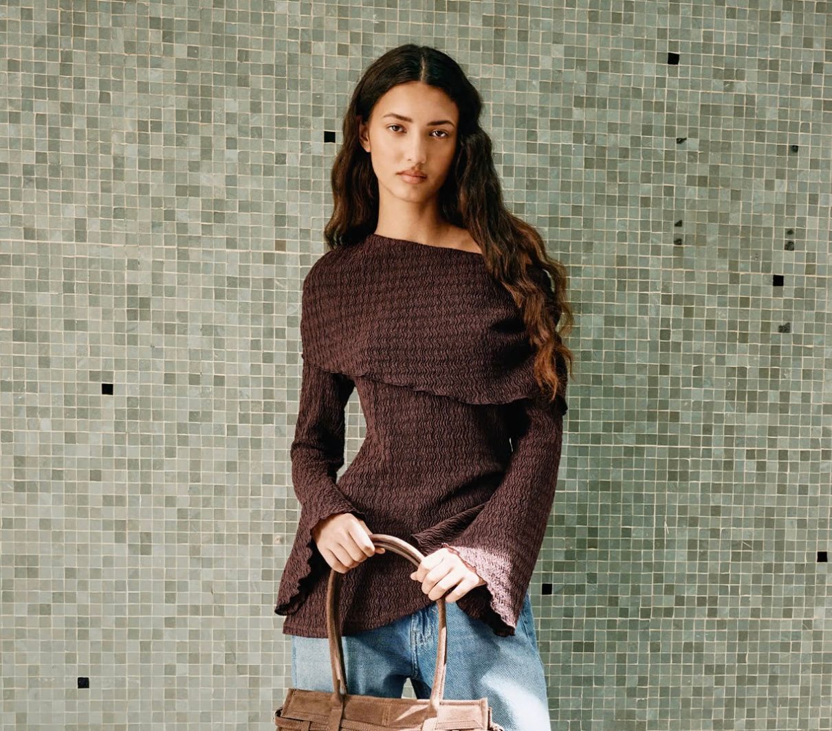 Model wearing a brown textured off-the-shoulder top with flared sleeves and light-wash jeans, holding a tan suede top-handle bag against a green mosaic tile wall