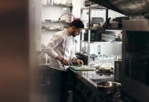 Chef in a white chef's jacket chopping ingredients on a board in a stainless steel restaurant kitchen