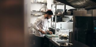 Chef in a white chef's jacket chopping ingredients on a board in a stainless steel restaurant kitchen