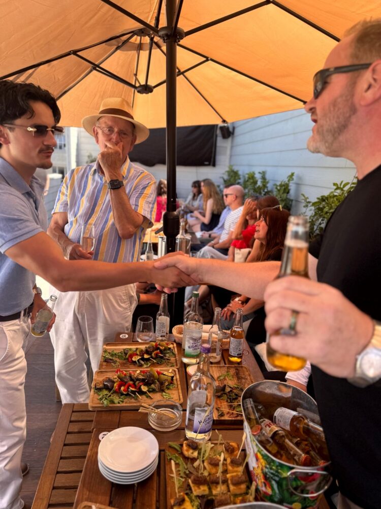 Guests shaking hands beside a table of food and drinks under a parasol at the SDEA Summer Social, July 2026