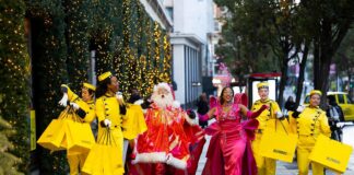 Selfridges Bellhops in yellow uniforms carrying yellow shopping bags walk along Oxford Street with Santa and the Showgirl