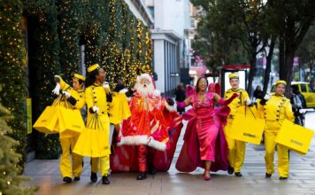 Selfridges Bellhops in yellow uniforms carrying yellow shopping bags walk along Oxford Street with Santa and the Showgirl