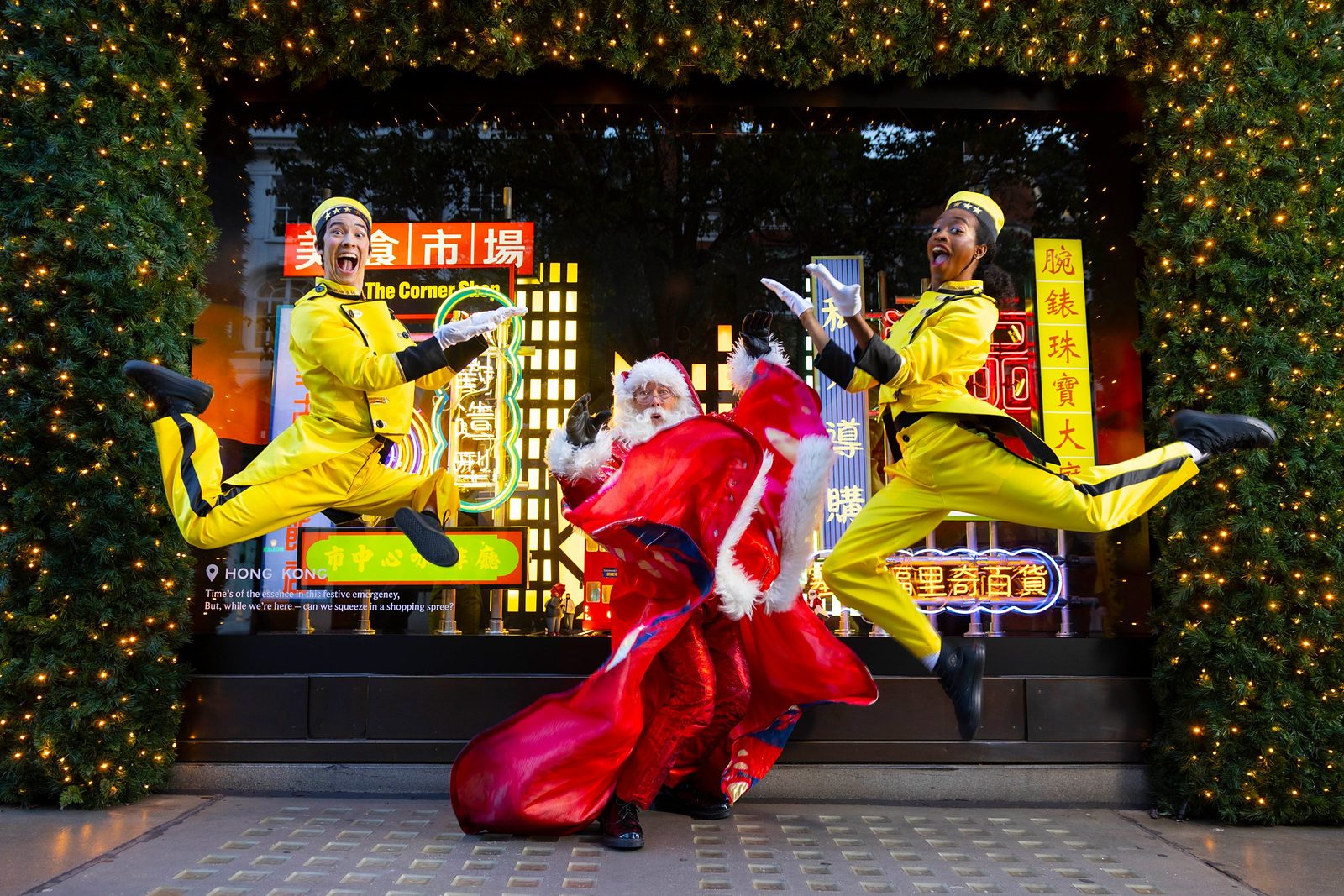Two Selfridges Bellhops in yellow uniforms leap either side of Santa in front of the Hong Kong window