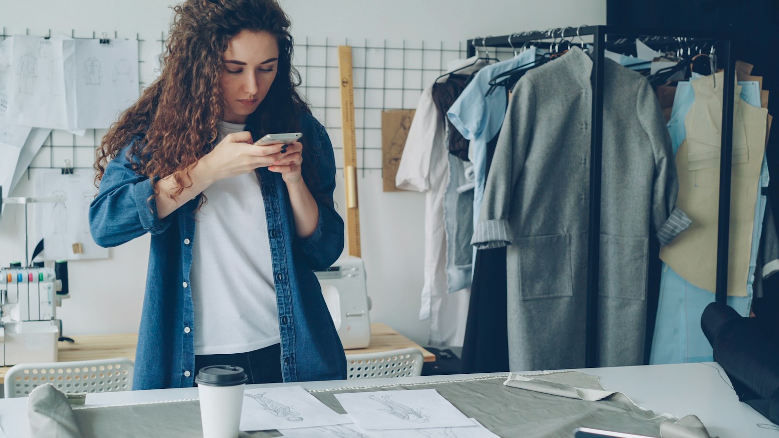 Woman using a smartphone to photograph clothes in a fashion studio, beside a rail of garments and a worktable of sketches